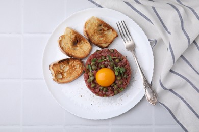 Photo of Tasty beef steak tartare served with yolk, capers, toasted bread and greens on white tiled table, top view