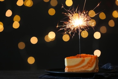 Photo of Cake with burning sparkler on black table against blurred festive lights. Space for text