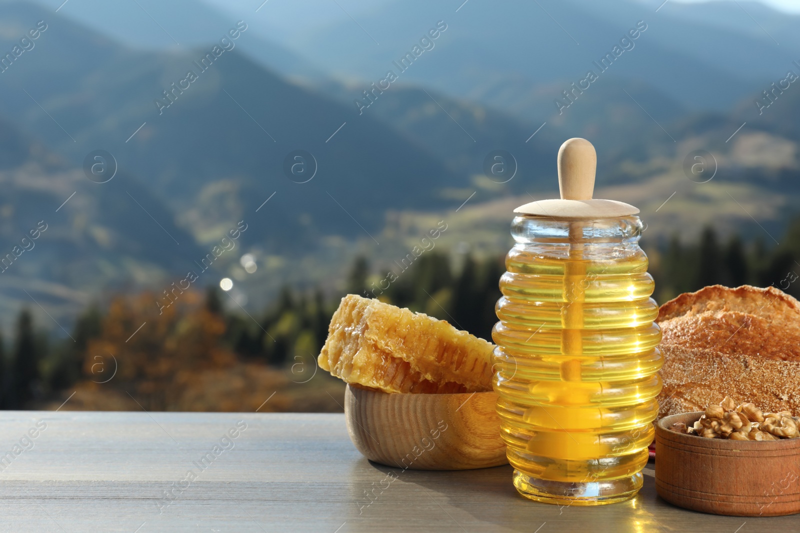 Photo of Fresh aromatic honey, combs, bread and nuts on grey wooden table against mountain landscape. Space for text