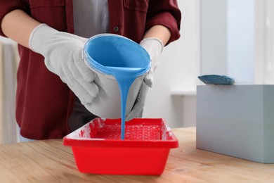 Photo of Woman pouring light blue paint from bucket into tray at wooden table indoors, closeup