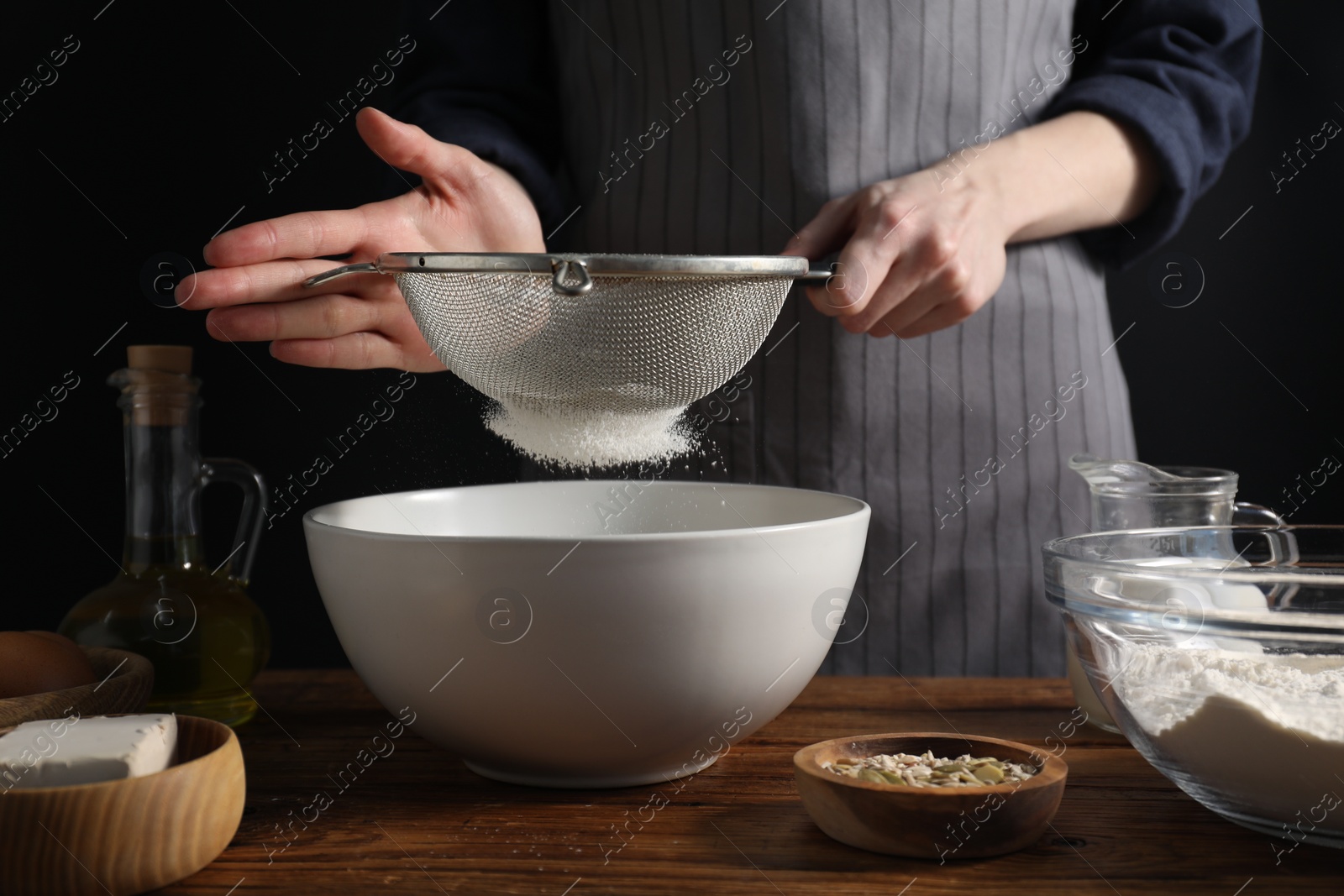 Photo of Making bread. Woman sifting flour over bowl at wooden table on dark background, closeup