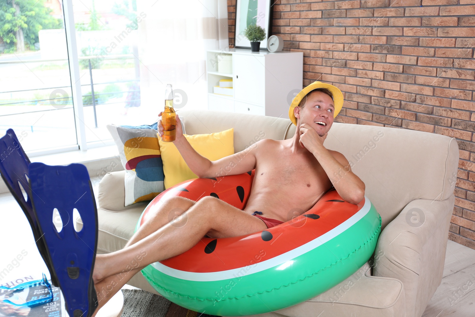 Photo of Shirtless man with inflatable ring and bottle of drink wearing flippers on sofa at home