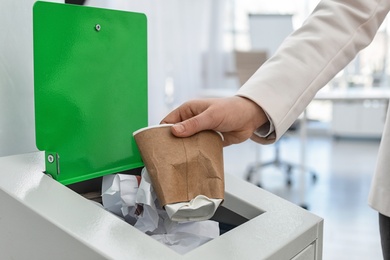 Photo of Woman putting used paper cup into trash bin in modern office, closeup. Waste recycling