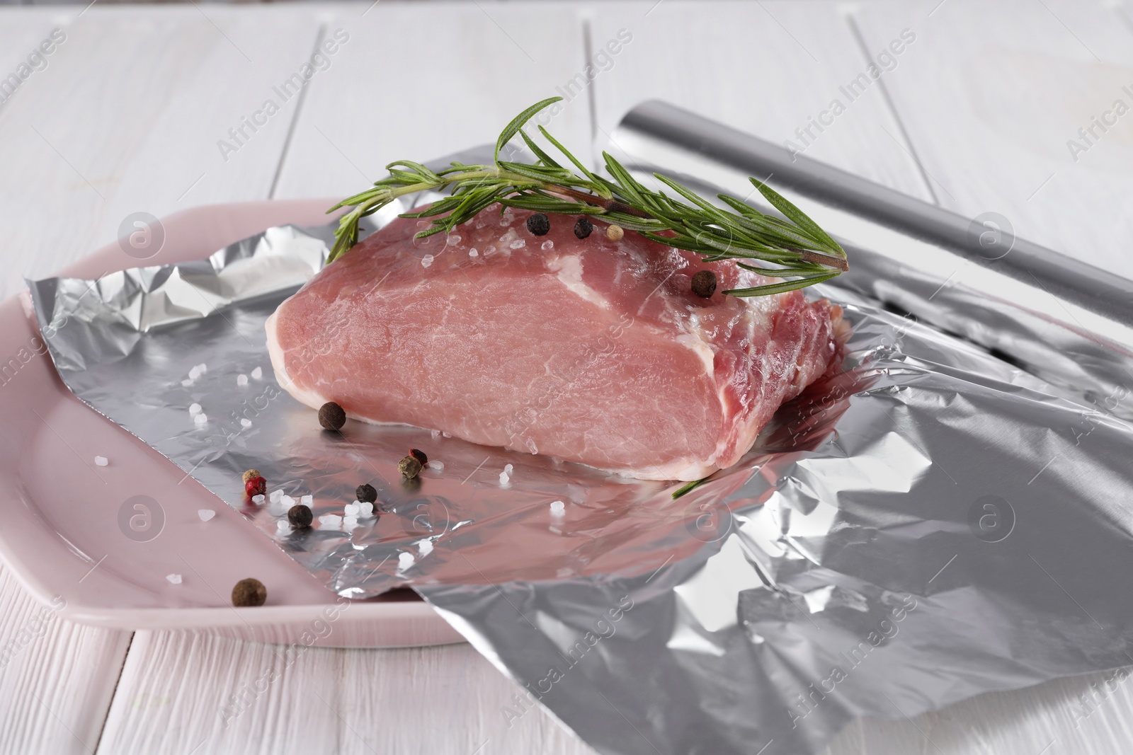 Photo of Aluminum foil with raw meat, rosemary and spices on white wooden table, closeup