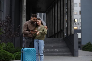 Photo of Being late. Worried couple with suitcase looking at watch near building outdoors, space for text