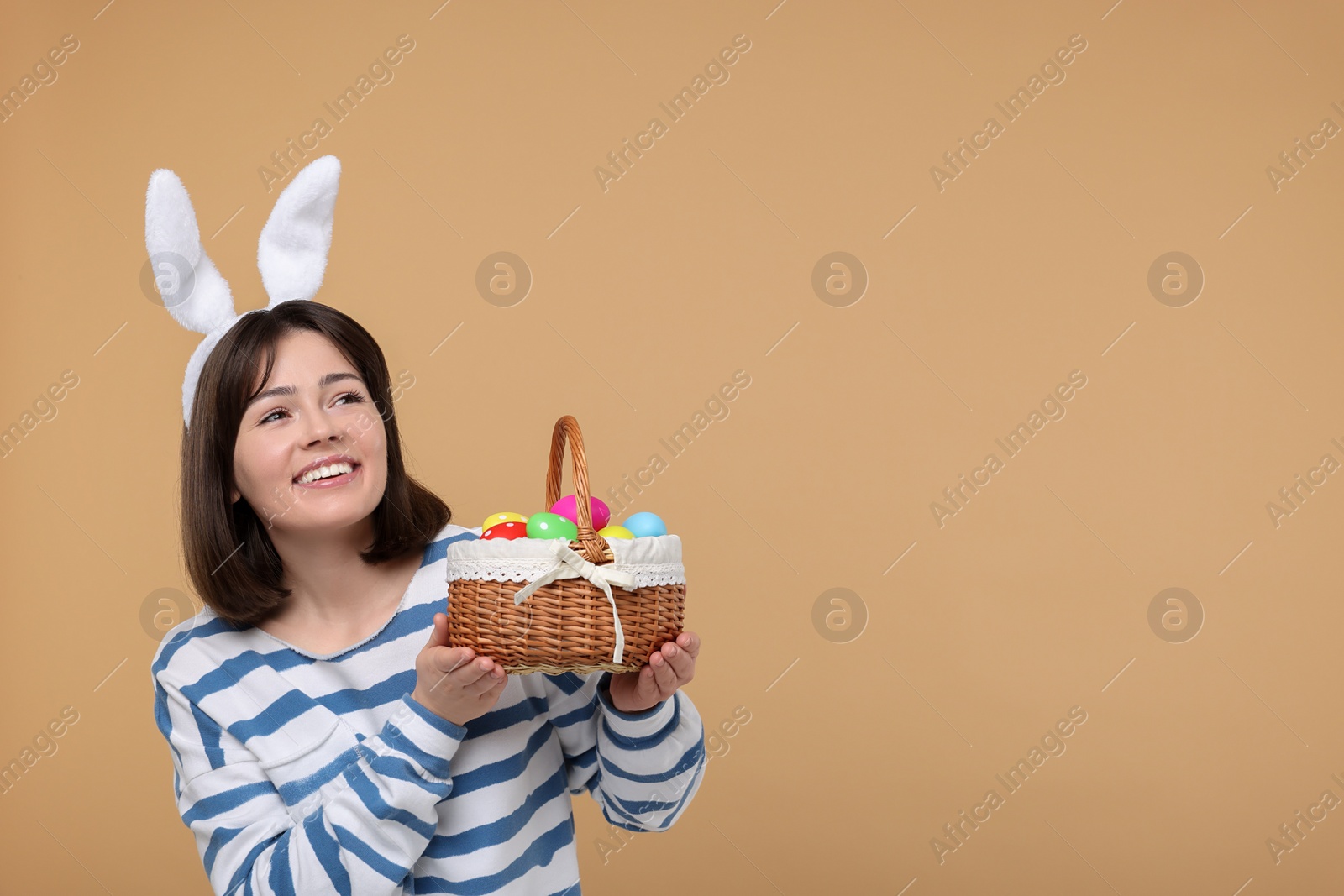 Photo of Easter celebration. Happy woman with bunny ears and wicker basket full of painted eggs on beige background, space for text