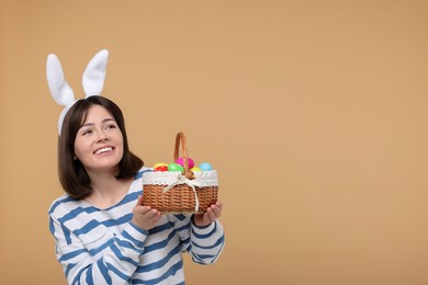 Photo of Easter celebration. Happy woman with bunny ears and wicker basket full of painted eggs on beige background, space for text