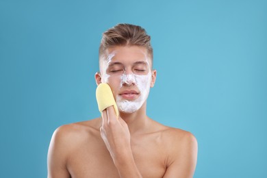 Photo of Young man washing off face mask with sponge on light blue background