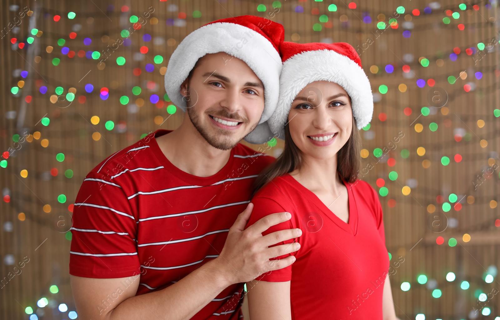 Photo of Young couple in Santa hats on blurred lights background. Christmas celebration