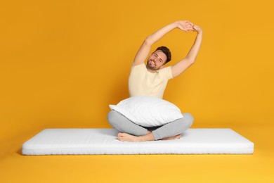 Photo of Man with pillow sitting on soft mattress and stretching against orange background