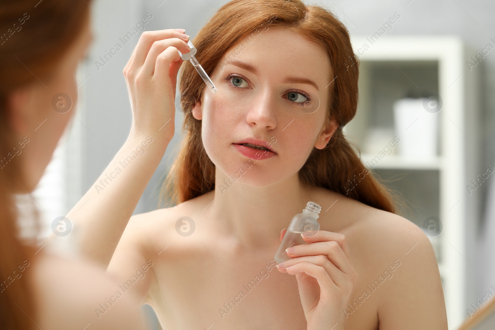 Photo of Beautiful woman with freckles applying cosmetic serum onto her face near mirror in bathroom