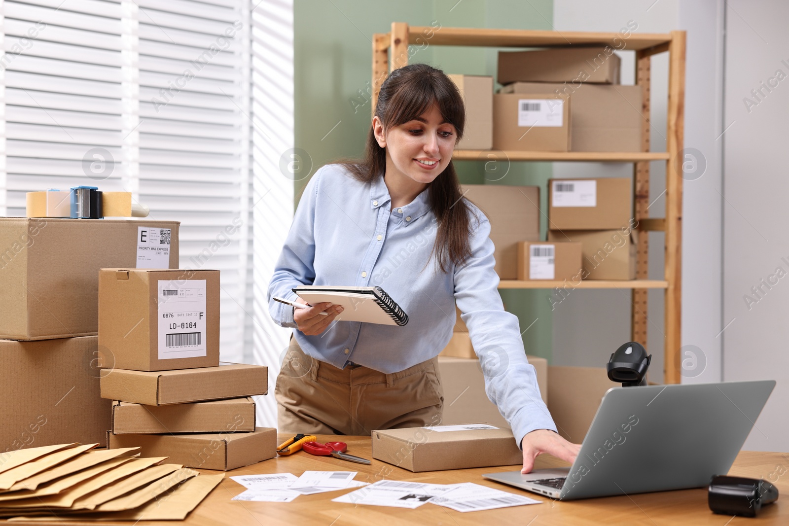 Photo of Parcel packing. Post office worker with notebook using laptop at wooden table indoors