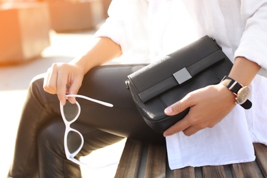 Photo of Young woman with stylish black bag and sunglasses on bench outdoors, closeup