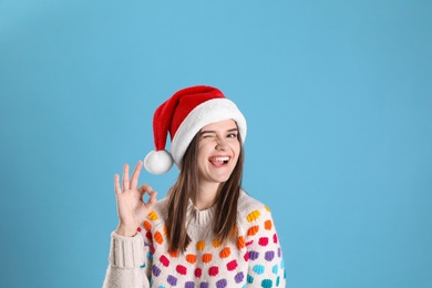 Pretty woman in Santa hat and festive sweater showing OK gesture on light blue background