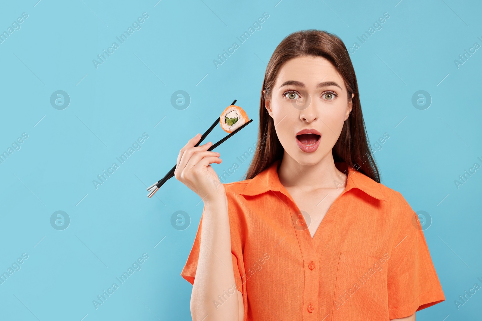 Photo of Emotional young woman holding sushi roll with chopsticks on light blue background. Space for text