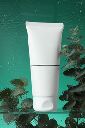 Tube with moisturizing cream and eucalyptus branches on green background, view through wet glass