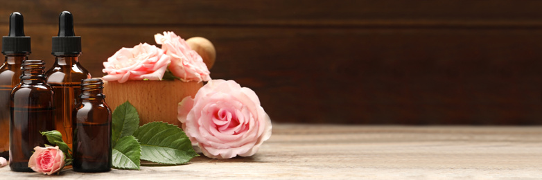 Bottles of rose essential oil and flowers on wooden table, space for text. Banner design