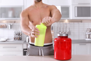 Photo of Young shirtless man holding protein shake in bottle near table with jar of powder at home, closeup
