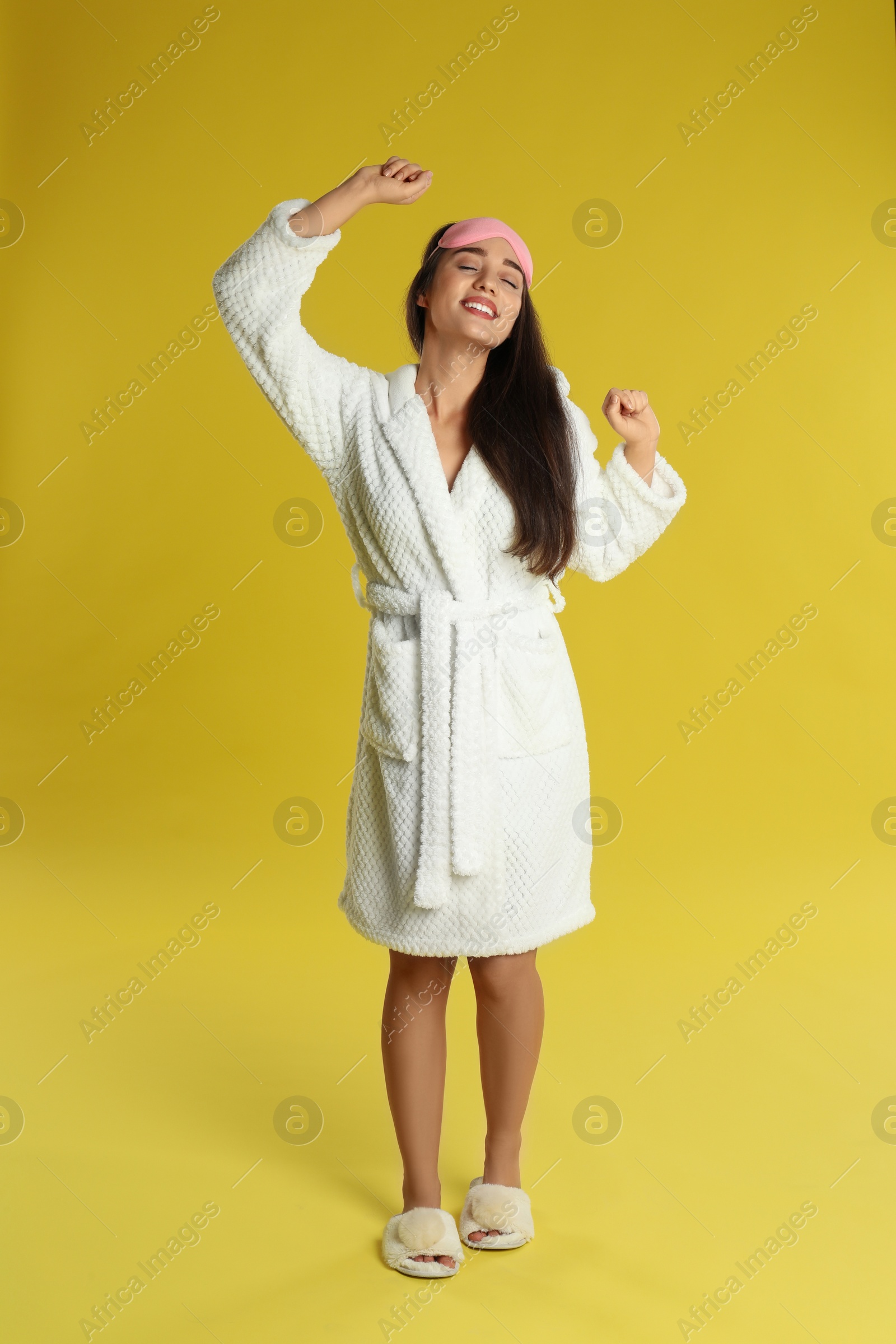 Photo of Beautiful young woman in bathrobe and eye sleeping mask stretching on yellow background