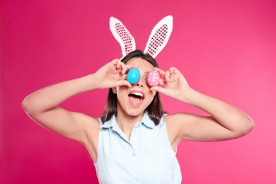 Beautiful woman in bunny ears headband holding Easter eggs near eyes on color background