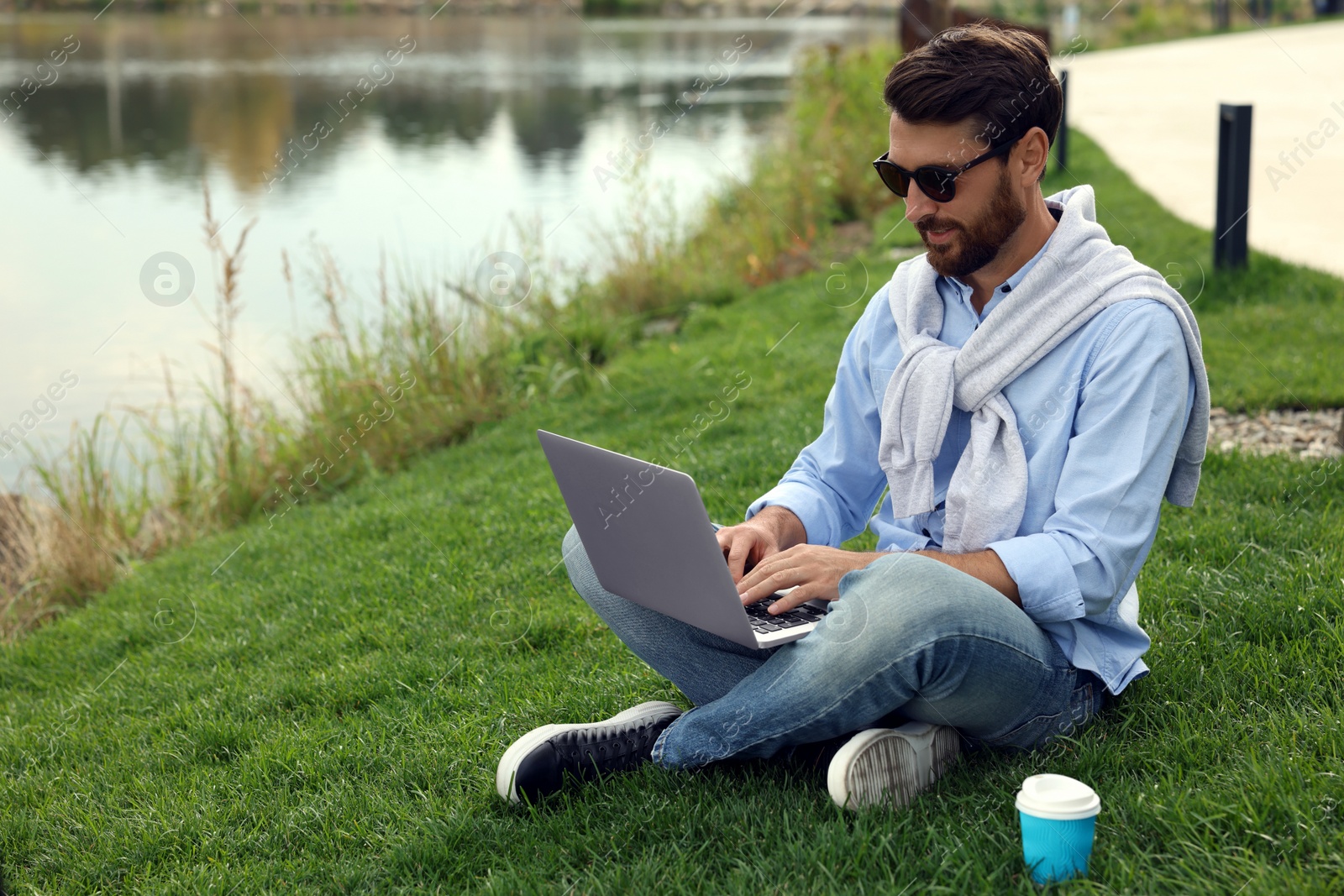 Photo of Man in sunglasses with laptop and coffee on green grass near lake