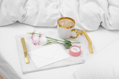 Tray with cup of coffee, flowers and beautiful engagement ring in box on white bed