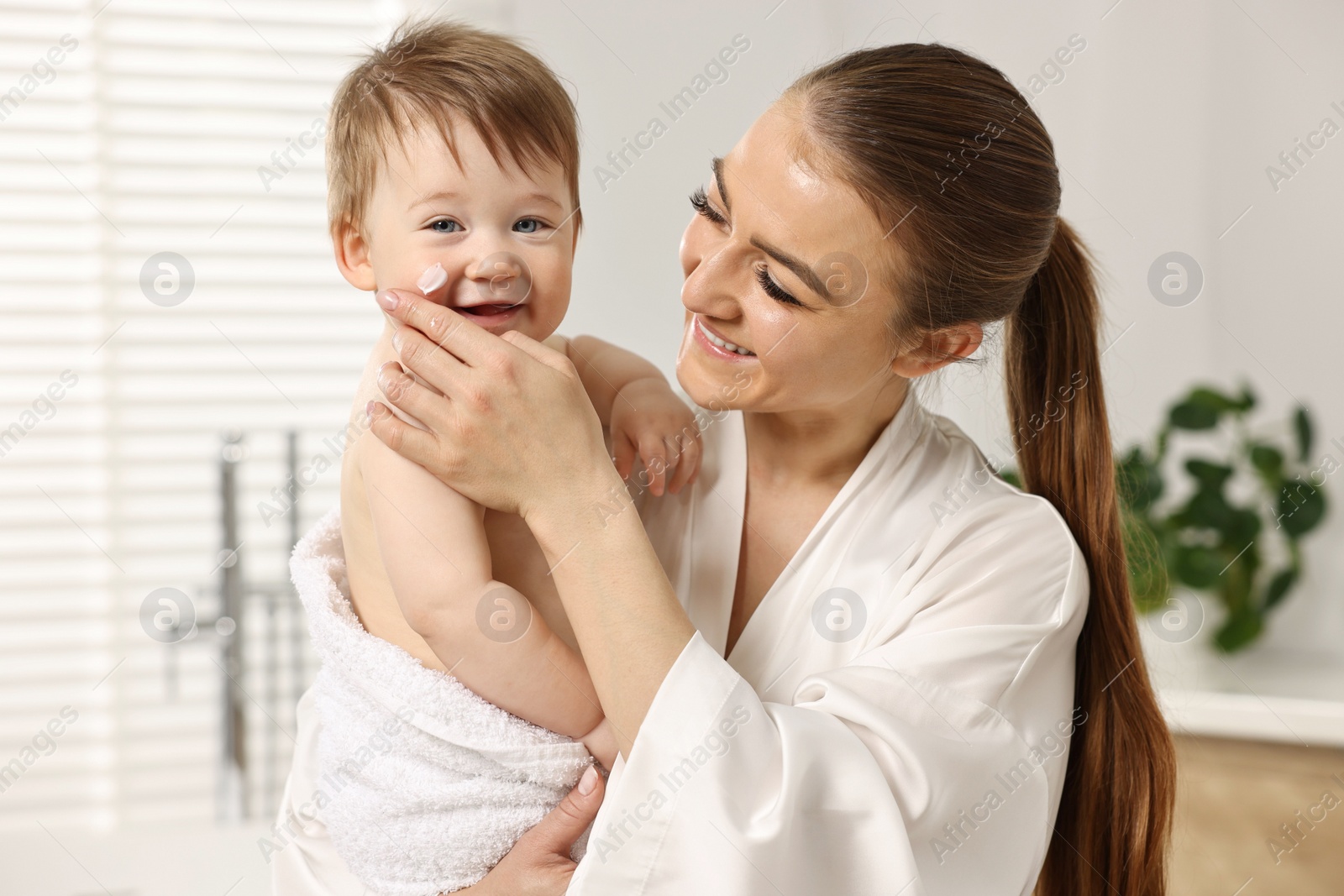 Photo of Happy mother applying moisturizing cream onto baby`s face in bathroom