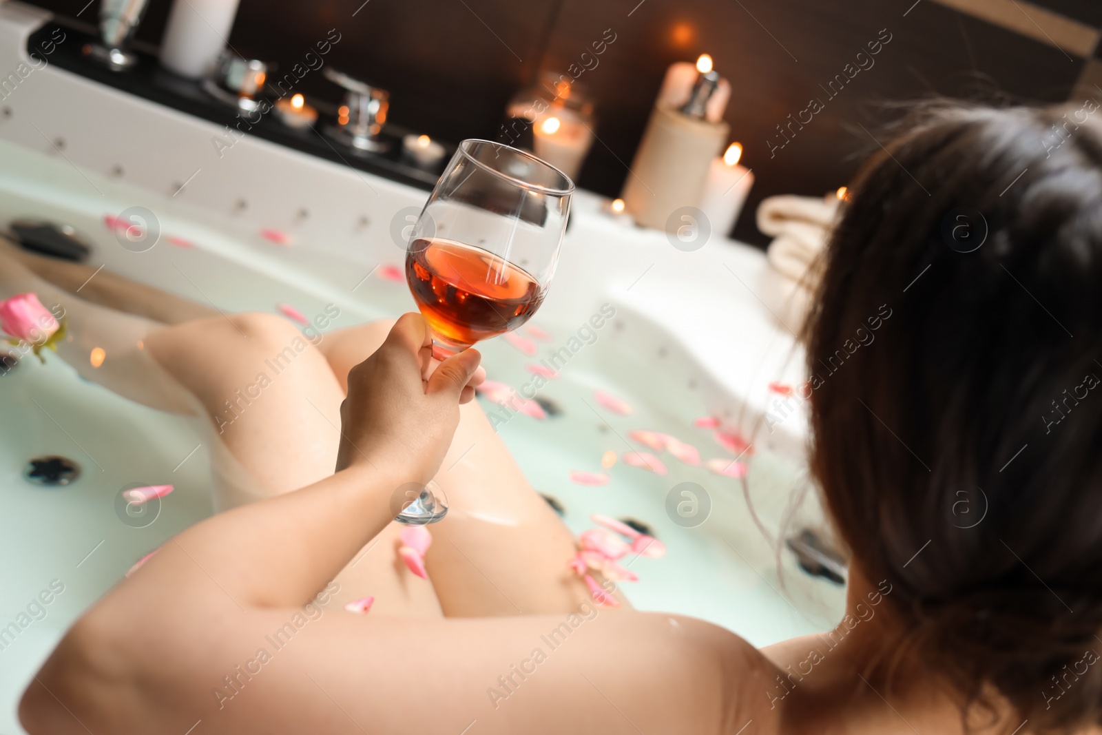 Photo of Woman holding glass of wine while taking bath with rose petals, back view. Romantic atmosphere