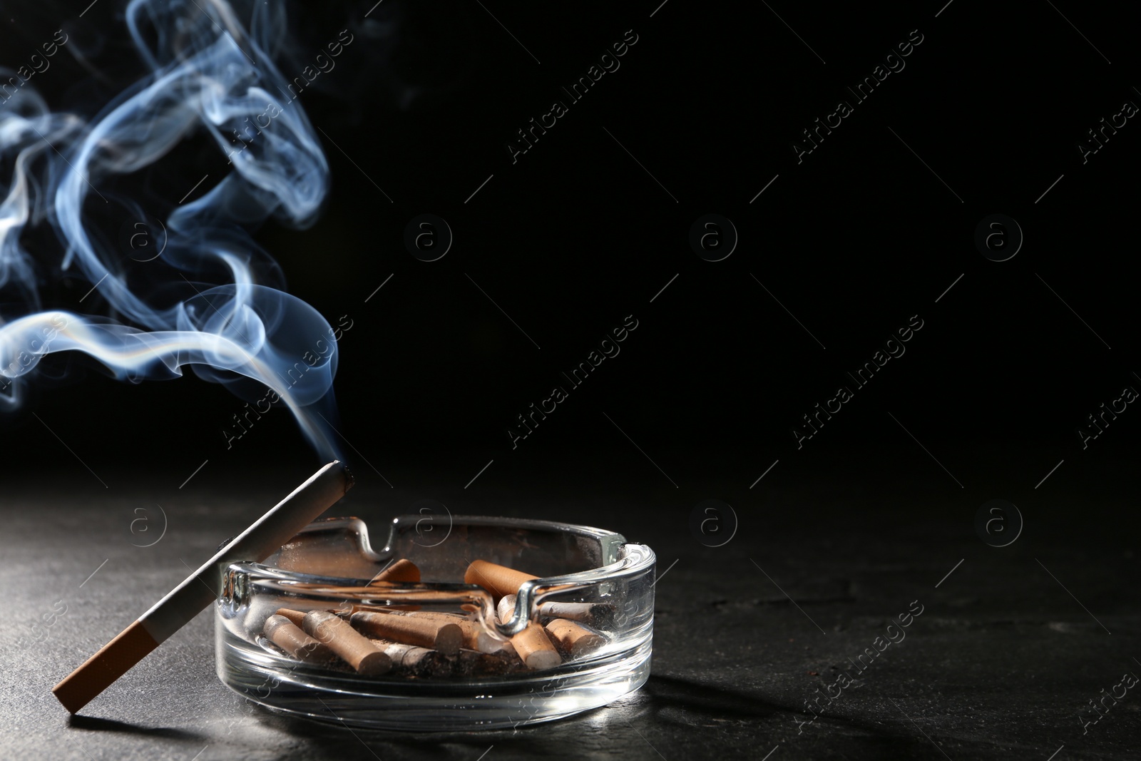 Photo of Glass ashtray with stubs and smoldering cigarette on grey table against black background. Space for text