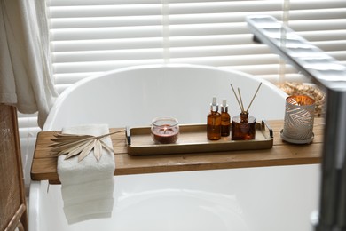 Photo of Wooden tray with cosmetic products, burning candles, reed air freshener and towel on bath tub in bathroom