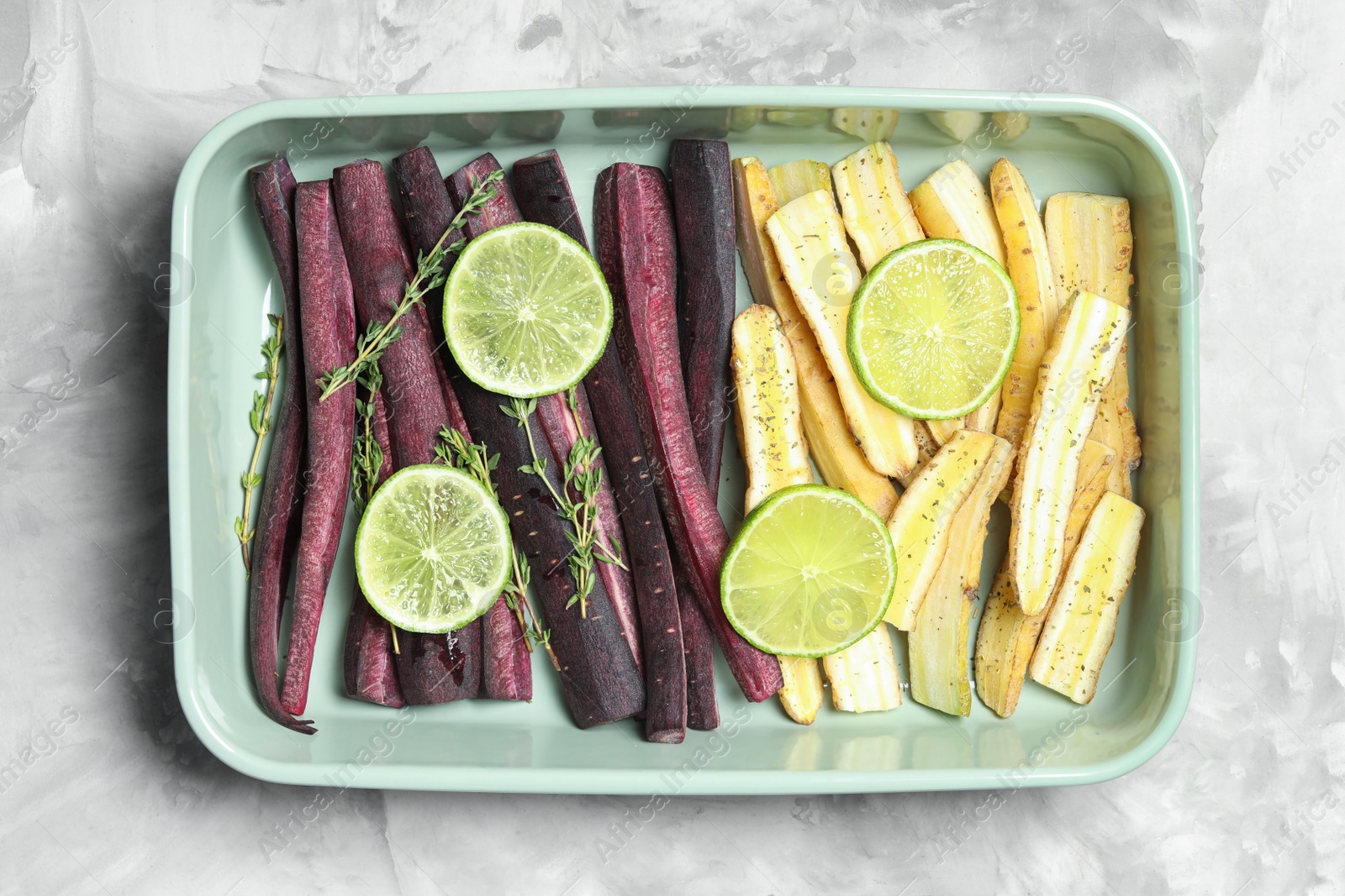 Photo of Raw black and white carrots with lime slices in baking dish on light grey table, top view