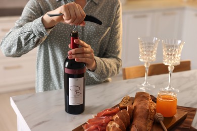 Romantic dinner. Woman opening wine bottle with corkscrew at table in kitchen, closeup