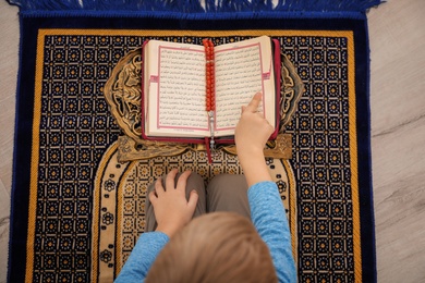 Little Muslim boy reading Koran on prayer rug indoors, top view