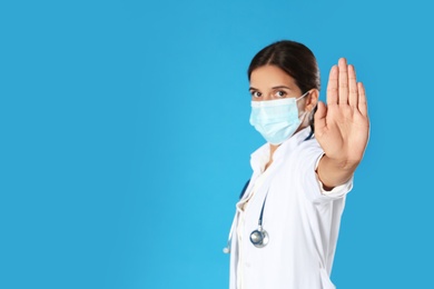 Photo of Doctor in protective mask showing stop gesture on blue background, space for text. Prevent spreading of coronavirus