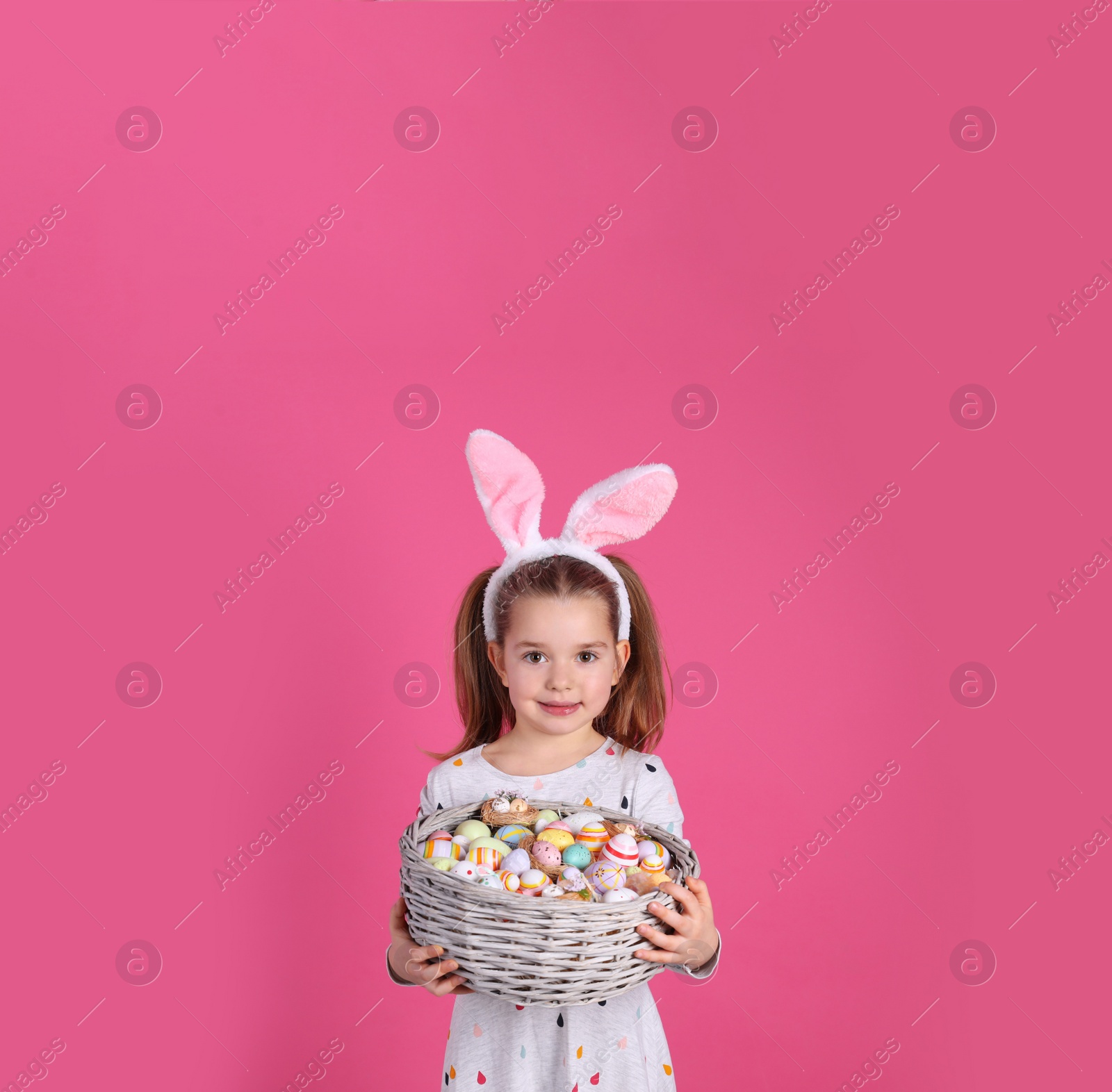 Photo of Adorable little girl with bunny ears holding wicker basket full of Easter eggs on pink background