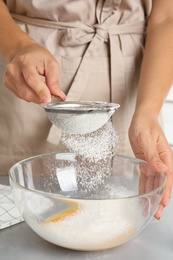 Woman sieving flour into bowl at light grey table, closeup. Cooking of delicious cake