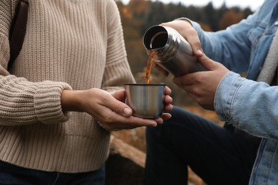 Photo of Boyfriend pouring hot drink from metallic thermos into cup lid for his girlfriend outdoors, closeup