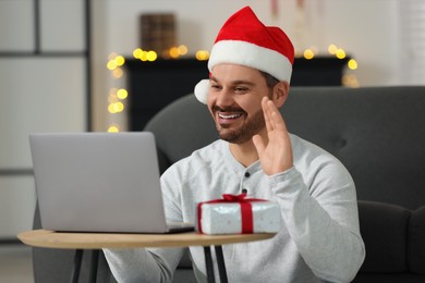 Photo of Celebrating Christmas online with exchanged by mail presents. Smiling man in Santa hat with gift waving hello during video call on laptop at home