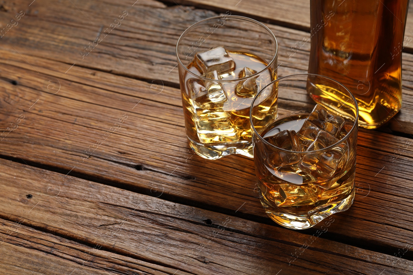 Photo of Whiskey with ice cubes in glasses and bottle on wooden table, closeup. Space for text