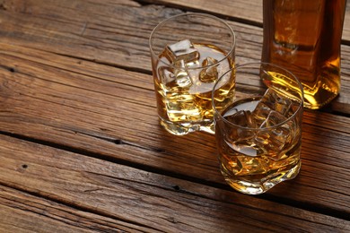 Whiskey with ice cubes in glasses and bottle on wooden table, closeup. Space for text