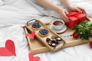 Photo of Tasty breakfast served in bed. Woman with gift box, tea, desserts, flowers and I Love You card at home, closeup