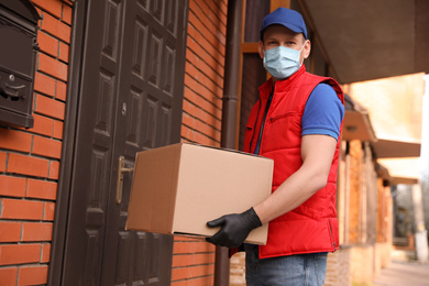 Photo of Courier in protective mask and gloves with box near house entrance. Delivery service during coronavirus quarantine