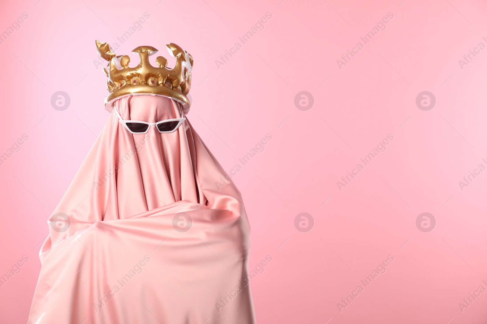 Photo of Glamorous ghost. Woman in sheet with sunglasses and crown on pink background, space for text