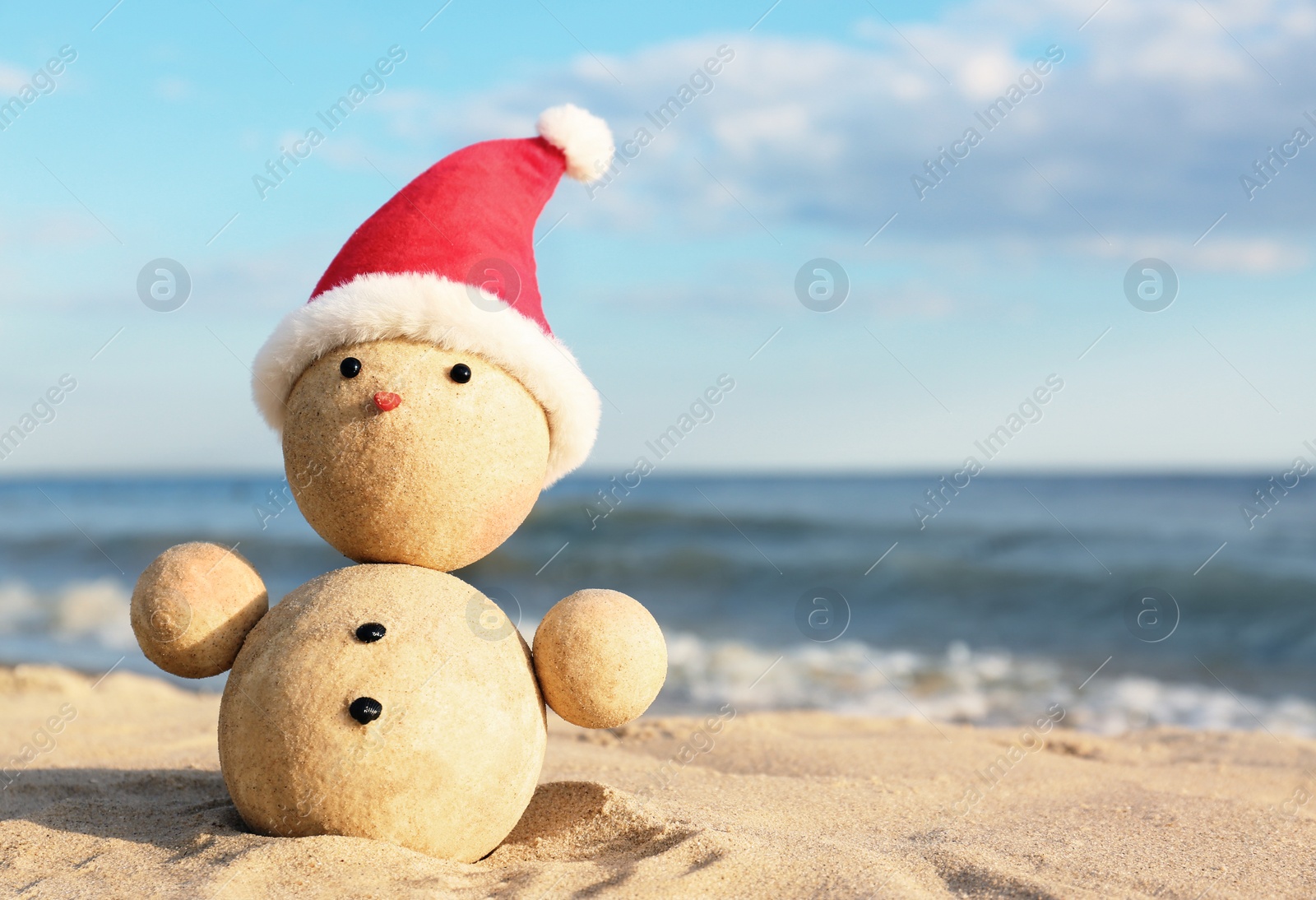 Photo of Snowman made of sand with Santa hat on beach near sea, space for text. Christmas vacation