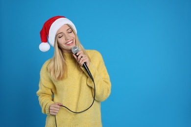 Photo of Happy woman in Santa Claus hat singing with microphone on blue background, space for text. Christmas music