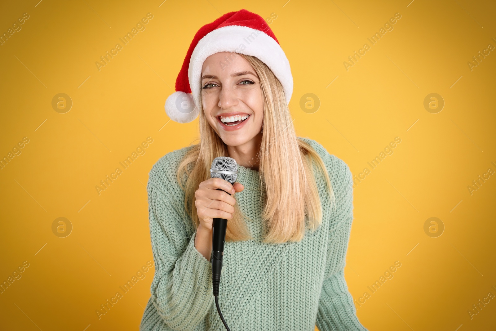 Photo of Happy woman in Santa Claus hat singing with microphone on yellow background. Christmas music