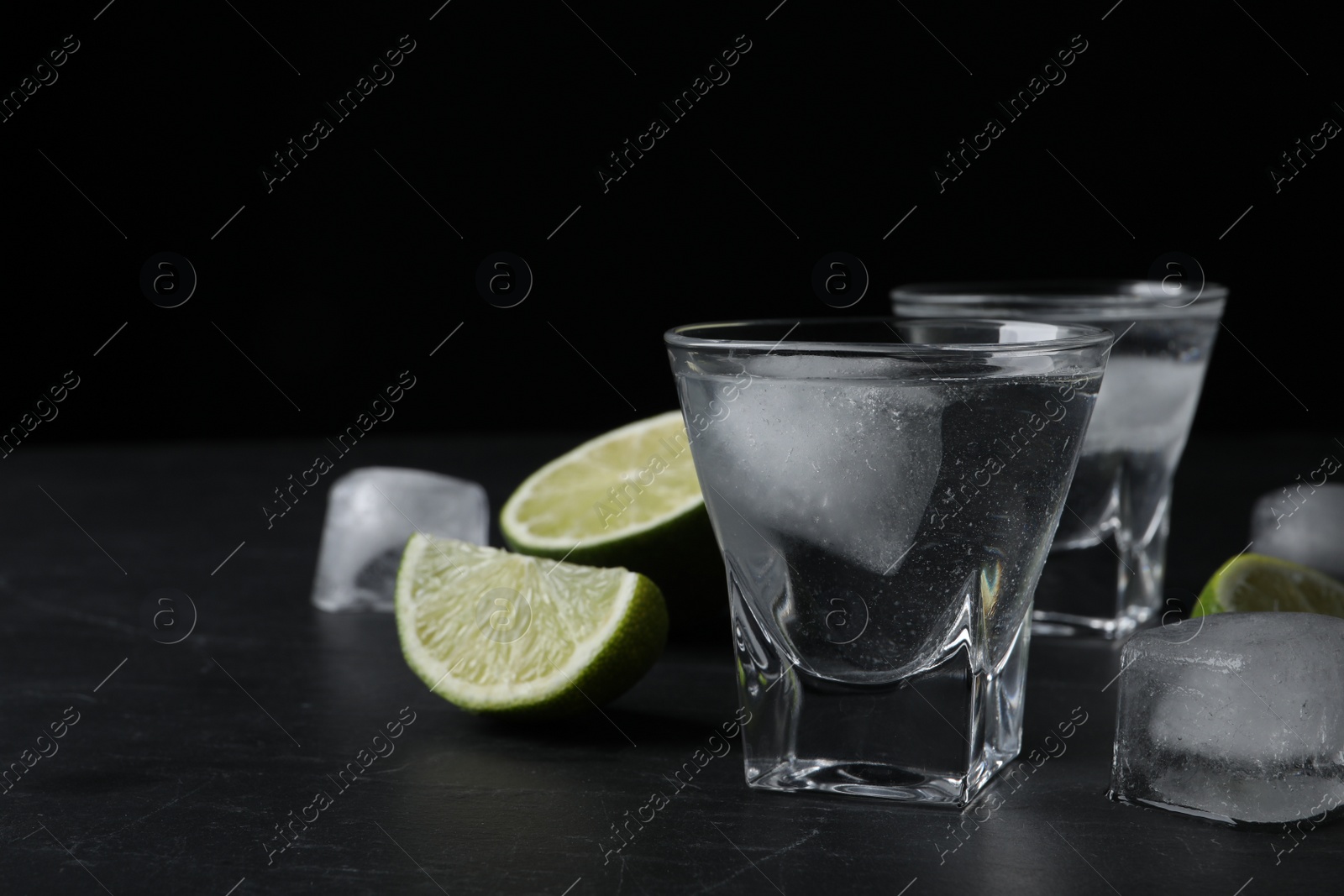 Photo of Shot glasses of vodka with lime slices and ice on black table. Space for text