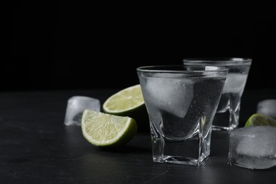 Photo of Shot glasses of vodka with lime slices and ice on black table. Space for text