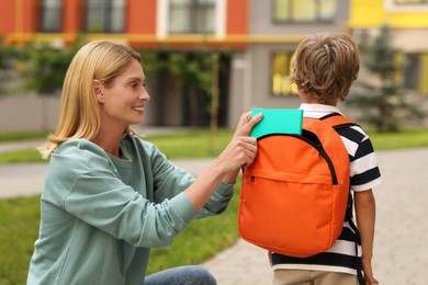 Photo of Happy woman putting notebook into with her son's backpack near kindergarten outdoors