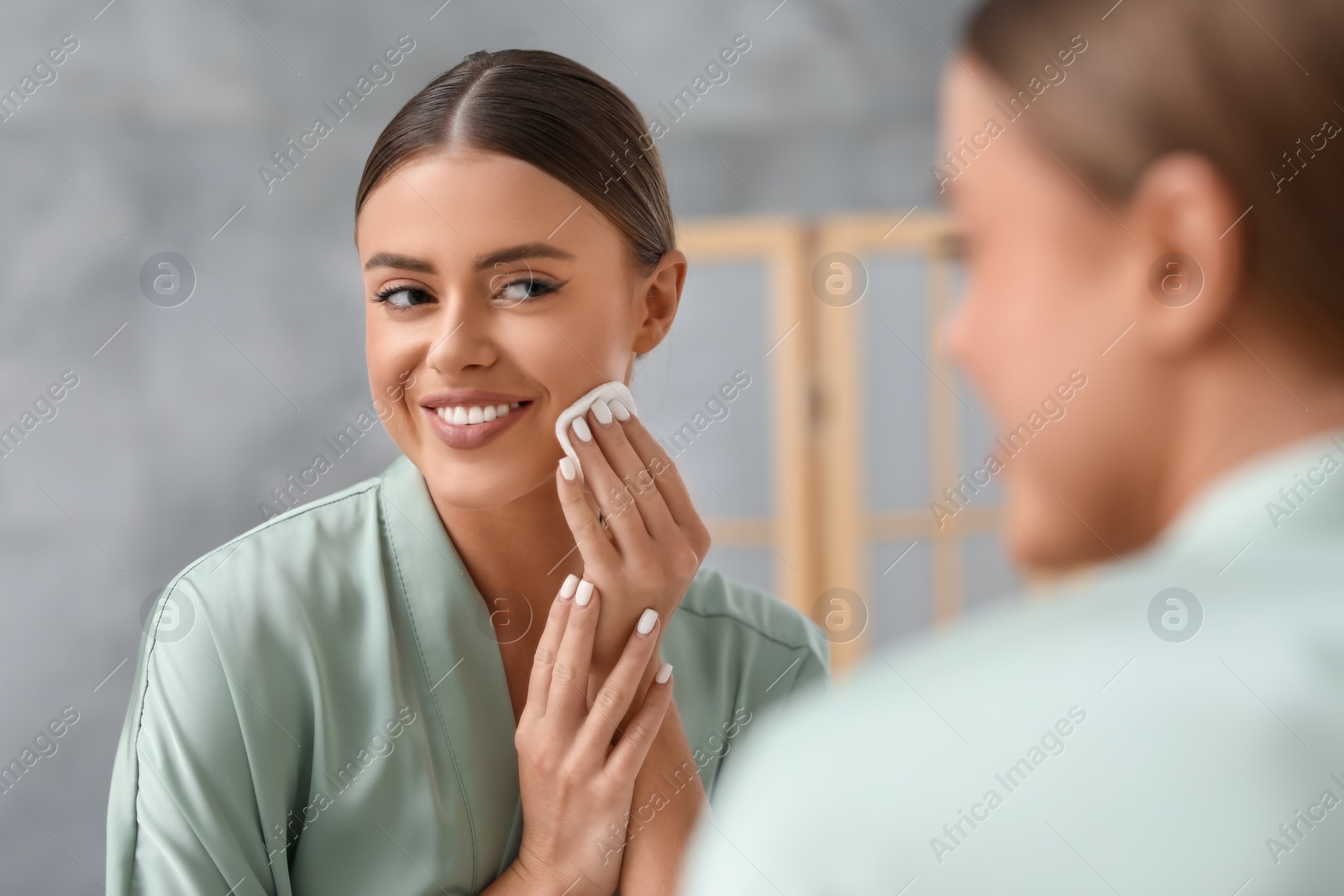 Photo of Beautiful woman removing makeup with cotton pad near mirror indoors